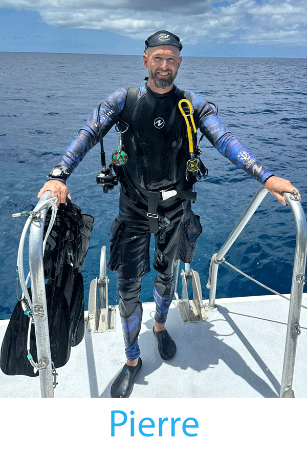 Man in wet suit holding fins, standing on boat with ocean in background.